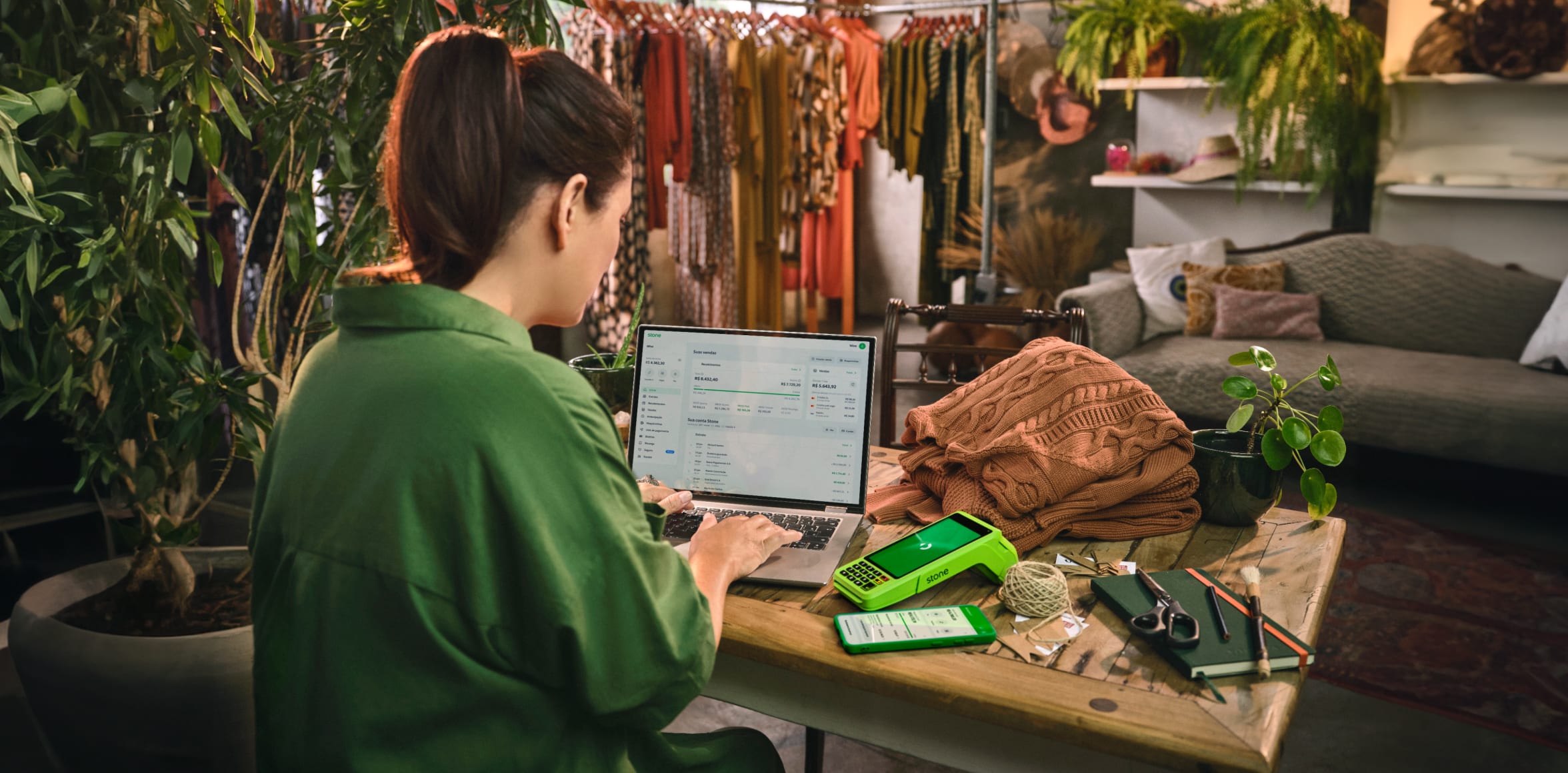 Mulher sentada e trabalhando no seu notebook e em cima da mesa contém maquininha de cartão Stone, caderno, tesoura, barbante, lápis, blusa e vazo de plantas