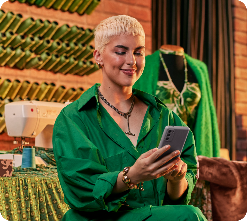 Empreendedora em sua loja de roupas sentada e utilizando o seu celular. Ao fundo uma máquina de costura, um manequim e linhas de costura verde penduradas na parede