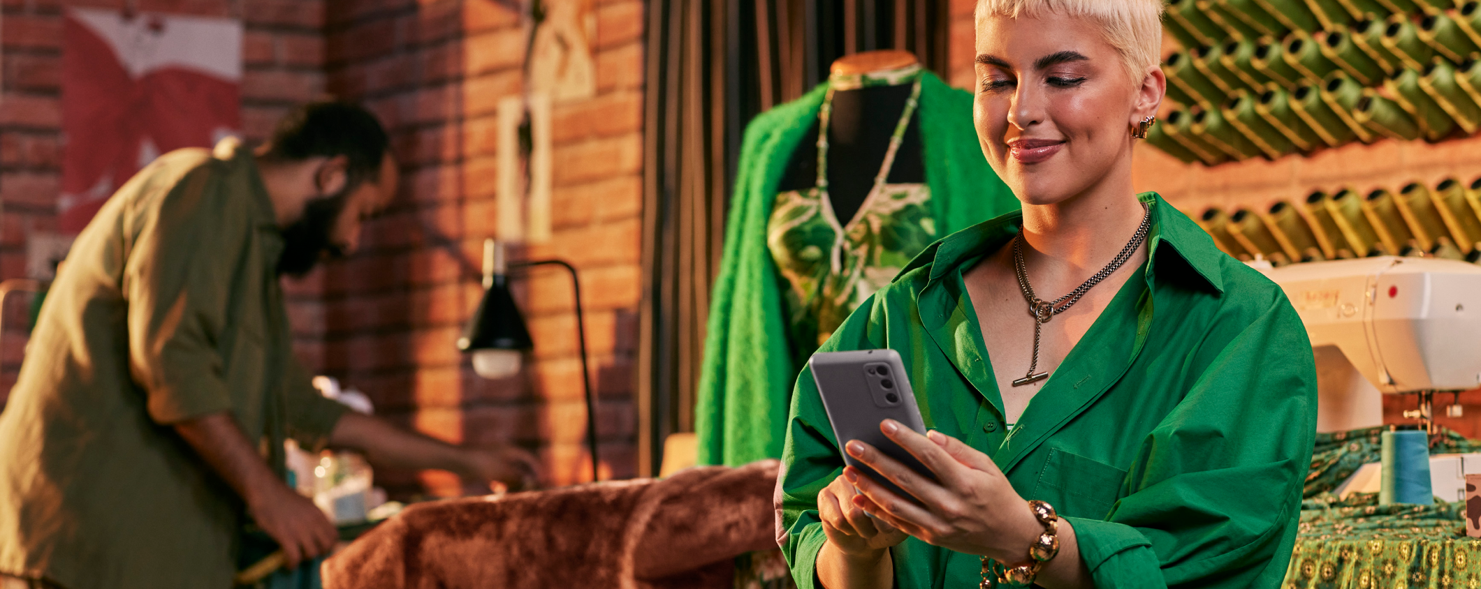 Mulher sorrindo, segurando um cartão Stone verde e um celular verde, em um ambiente de loja ou escritório.