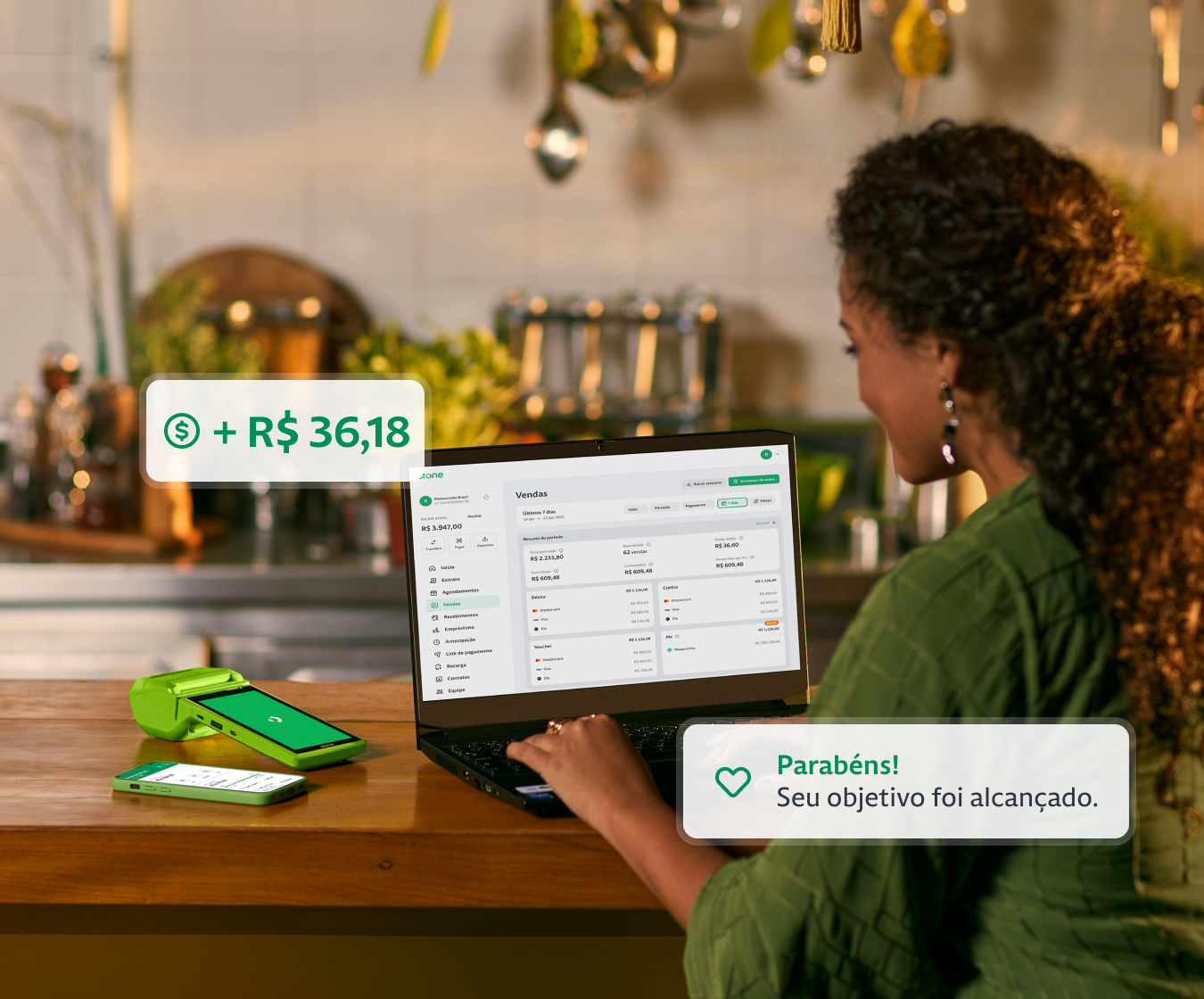 Empreendedora utilizando o seu notebook na bancada da cozinha do seu restaurante. Ao lado do notebook, constam uma máquina de cartão e um celular. 