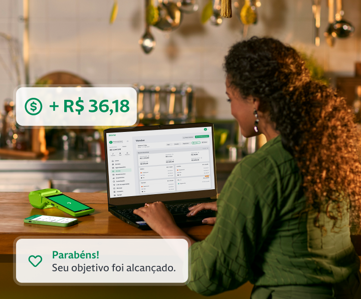 Empreendedora utilizando o seu notebook na bancada da cozinha do seu restaurante. Ao lado do notebook, constam uma máquina de cartão e um celular. 