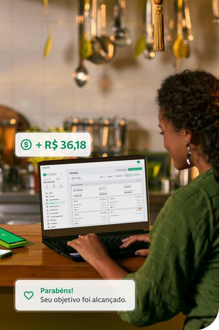 Empreendedora utilizando o seu notebook na bancada da cozinha do seu restaurante. Ao lado do notebook, constam uma máquina de cartão e um celular. 