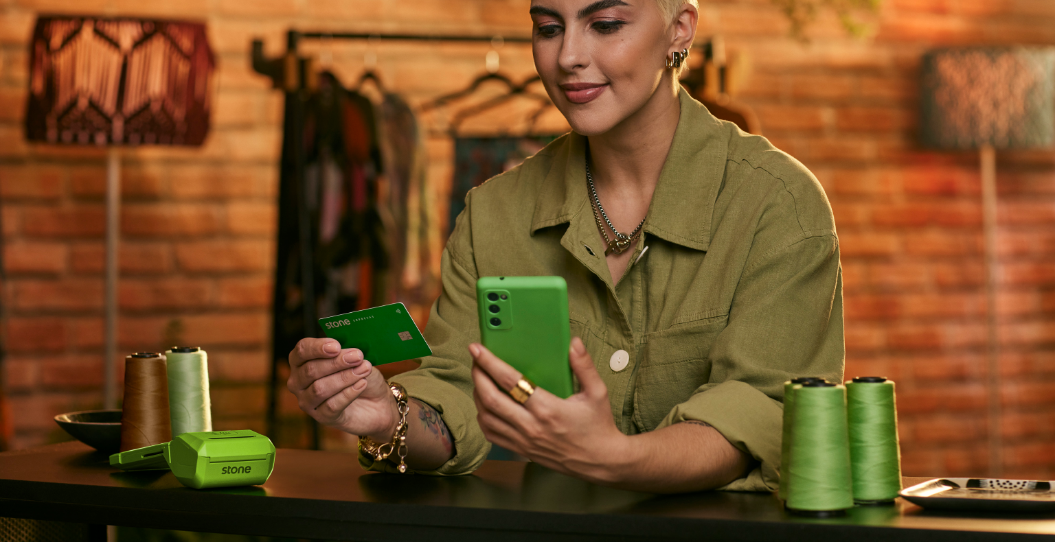 Empreendedora apoiada no balcão e segurando o seu celular com capinha verde em uma mão e na outra o cartão Stone