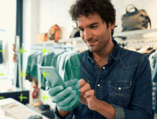 Empreendedor segurando o celular em sua loja de roupas. Atrás algumas araras com roupas penduradas e malas. 