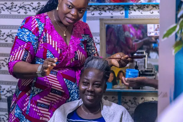 A stylist checking her tools while a customer looks on in a traditional hair salon, illustrating the sense of community and service in West African salons.