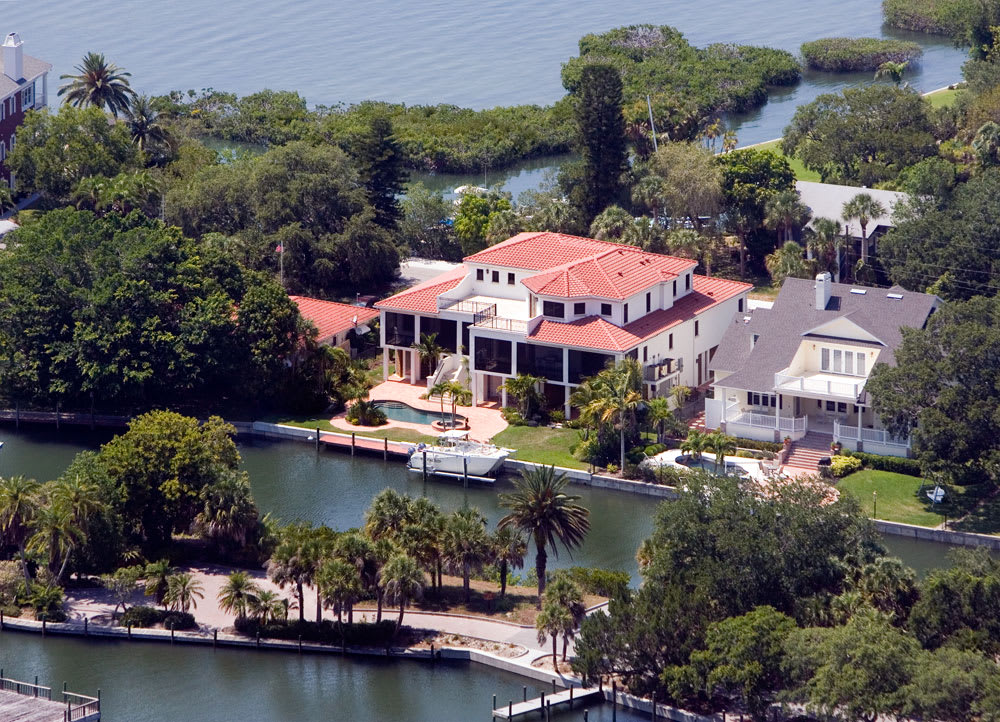Aerial view of a large, Mediterranean-style house with a red tile roof by a canal, surrounded by trees and greenery, with a dock and a boat in front, near a waterfront area.