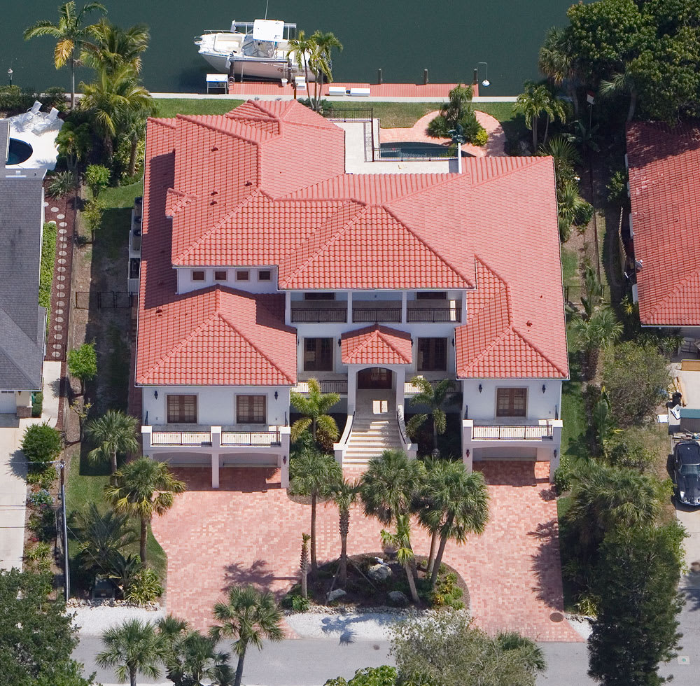 Aerial view of a large, two-story house with a red-tiled roof, spacious driveway, palm trees, and a backyard pool next to a canal with a docked white boat.