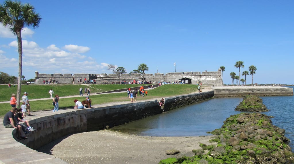 Visitors walk along a stone seawall and grassy area near a historic fort by the water under a bright blue sky with palm trees.