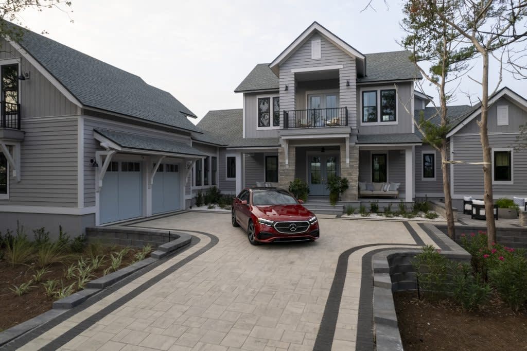 A red Mercedes-Benz is parked in the driveway of a large two-story gray house with white trim, a front balcony, and an attached garage. The landscaping features plants and neatly paved walkways.