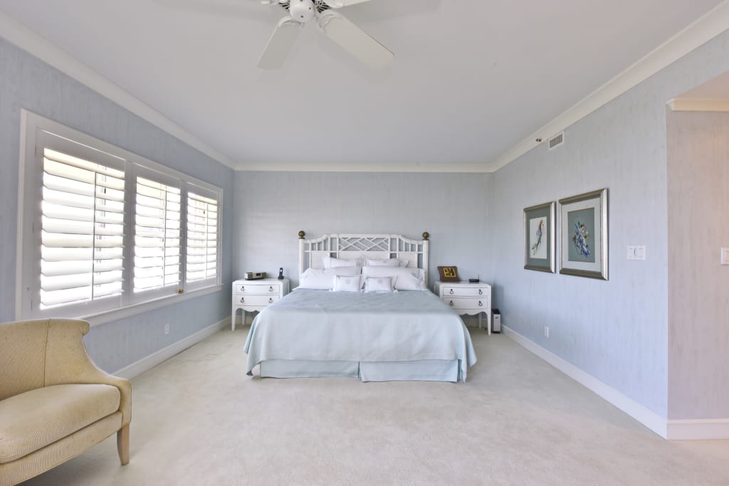 A bright, spacious bedroom with a large bed covered in light blue bedding, two white nightstands, framed art on the walls, a beige armchair, shuttered windows, and a ceiling fan.