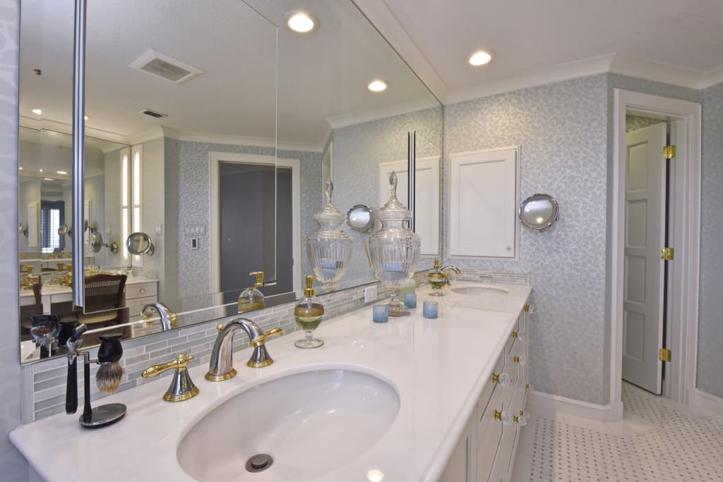 A spacious bathroom with a long white double sink vanity, gold fixtures, glass jars, light blue wallpaper, a large mirror, overhead lights, and an open door showing another room.