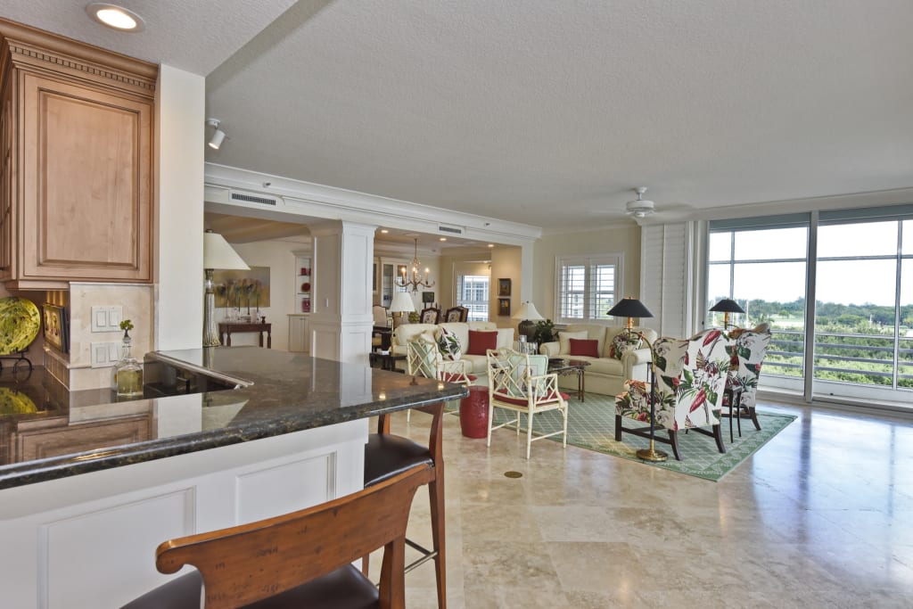 Open-plan living area with a kitchen counter in the foreground, barstool seating, a sitting area with floral armchairs, large windows offering a scenic view, and neutral-toned tile flooring throughout.