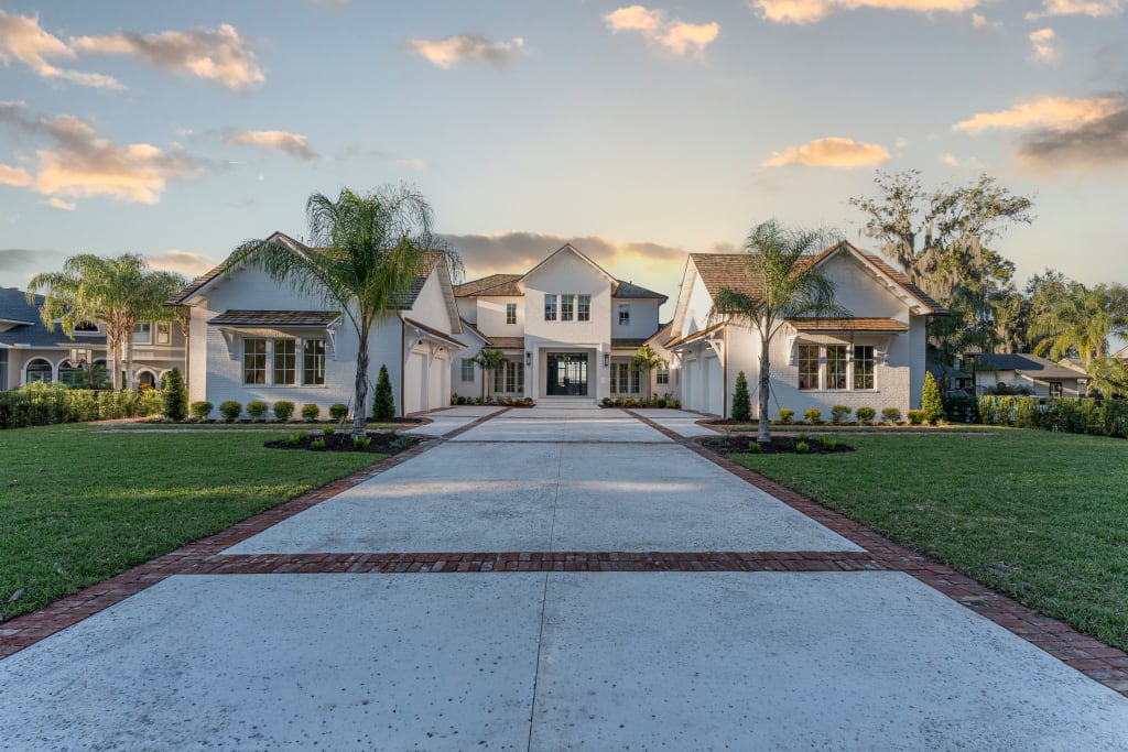 A large, elegant two-story house with white exterior, tall windows, and gabled roofs, sits at the end of a wide driveway lined with grass, palm trees, and manicured landscaping under a partly cloudy sky at sunset.