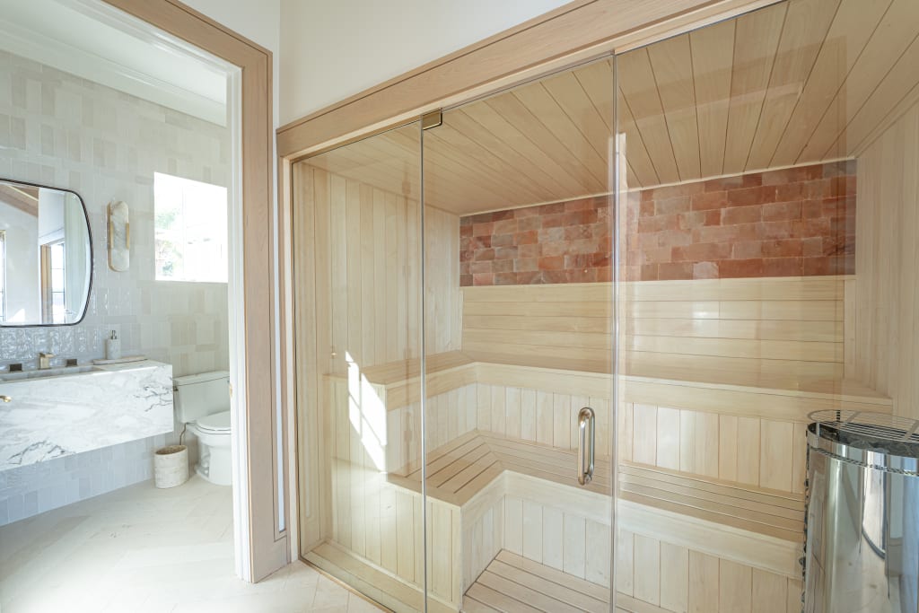 A modern bathroom with a marble sink, toilet, and a large glass-enclosed wooden sauna featuring a tiered bench and a wall of salt blocks.