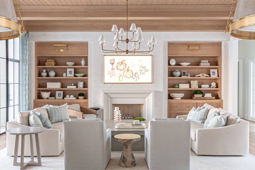 Bright, elegant living room with neutral tones, four cushioned chairs around a round table, built-in wooden shelves with decor, a white fireplace, and a framed fall-themed art piece above the mantle.