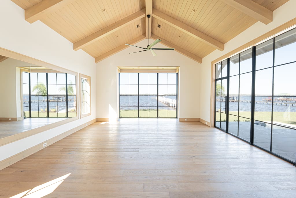 Spacious, empty room with light wood floors, vaulted wood ceiling, large windows, and glass doors offering views of water and a dock; a large wall mirror reflects the bright, airy space.