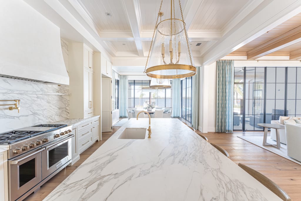 Bright, modern kitchen with a marble island, gold pendant lights, a gas stove with a marble backsplash, white cabinetry, and large windows letting in natural light. Open layout connects to a living area.
