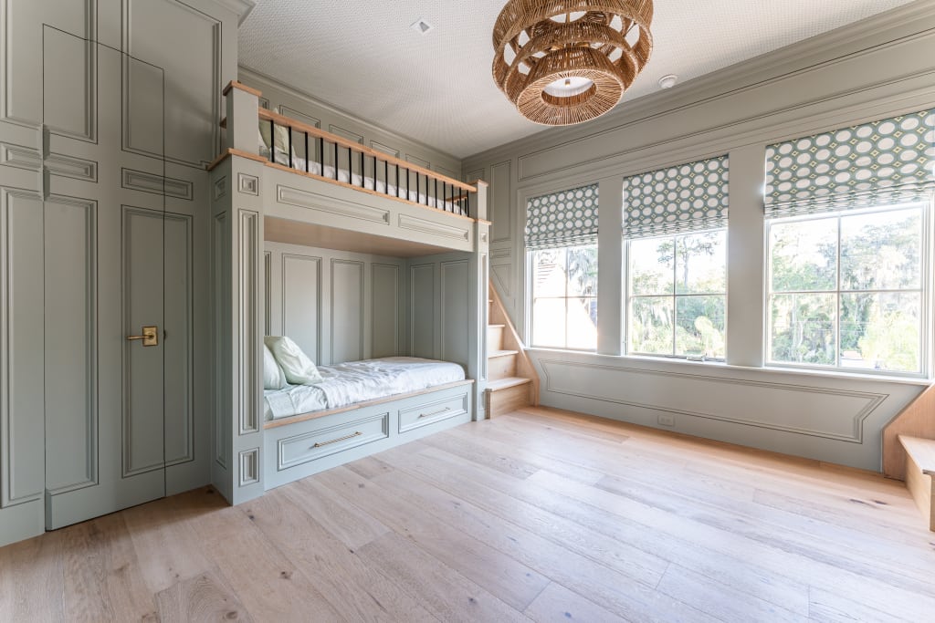 A spacious, light-filled bedroom with built-in light gray bunk beds, wooden steps, large windows with patterned shades, wood flooring, and a woven ceiling light fixture. The décor is modern and minimal.