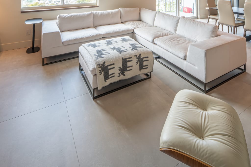 Modern living room with a large white sectional sofa, plush ottoman covered with a patterned blanket, a round side table, and a cushioned chair on a light-colored tile floor near large windows.