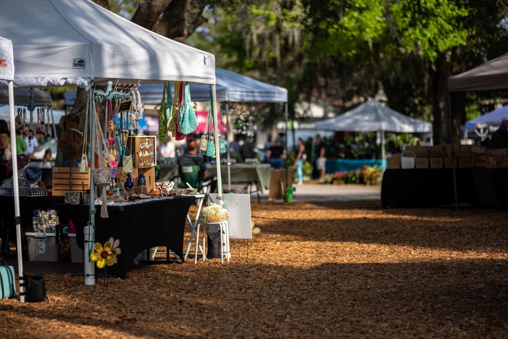 Outdoor market with white tents, tables displaying handmade crafts, jewelry, and bags. People browse the stalls under large trees, and sunlight filters through the leaves onto the woodchip-covered ground.