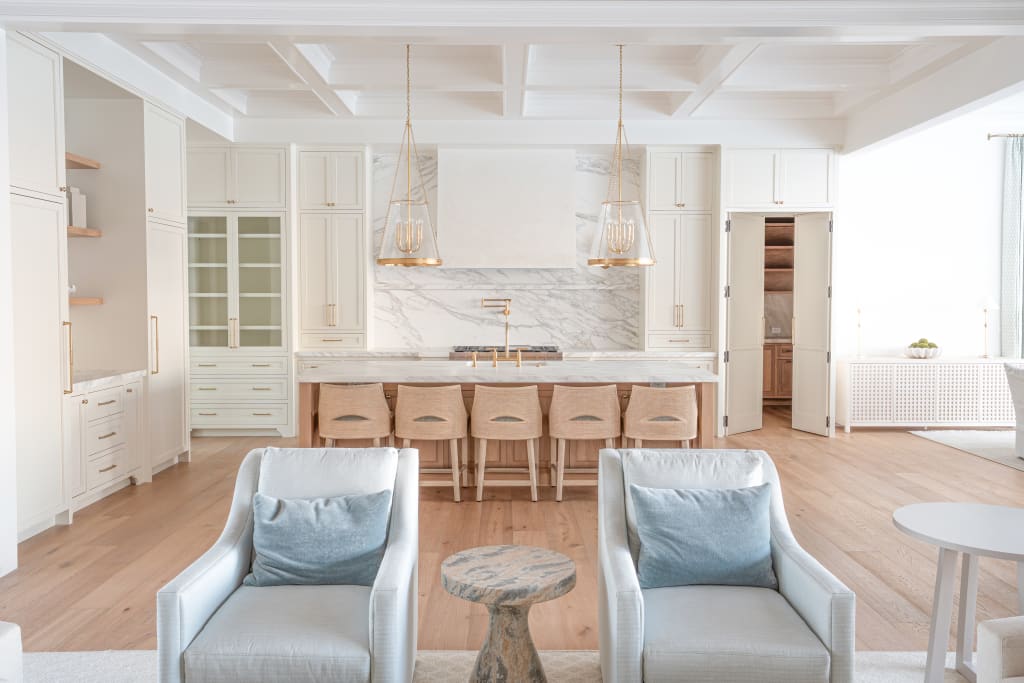 Modern, bright kitchen with marble backsplash, white cabinets, six wooden barstools at an island, two armchairs with blue pillows, a small round table, and pendant lights, all set on light wood flooring.