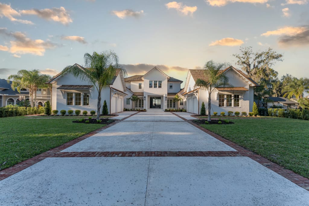 A large modern house with a white exterior, multiple peaked roofs, and large windows. A wide driveway lined with grass and small palm trees leads up to the main entrance. The sky is lightly clouded at sunset.