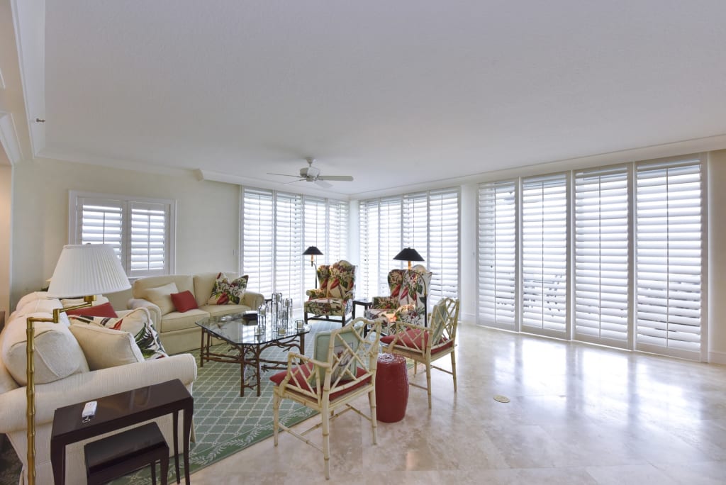 Bright living room with large windows covered by white shutters, light-colored walls and floor, tropical-patterned chairs, a beige sofa, glass coffee table, and green area rug. Lamps and decorative pillows add a cozy touch.