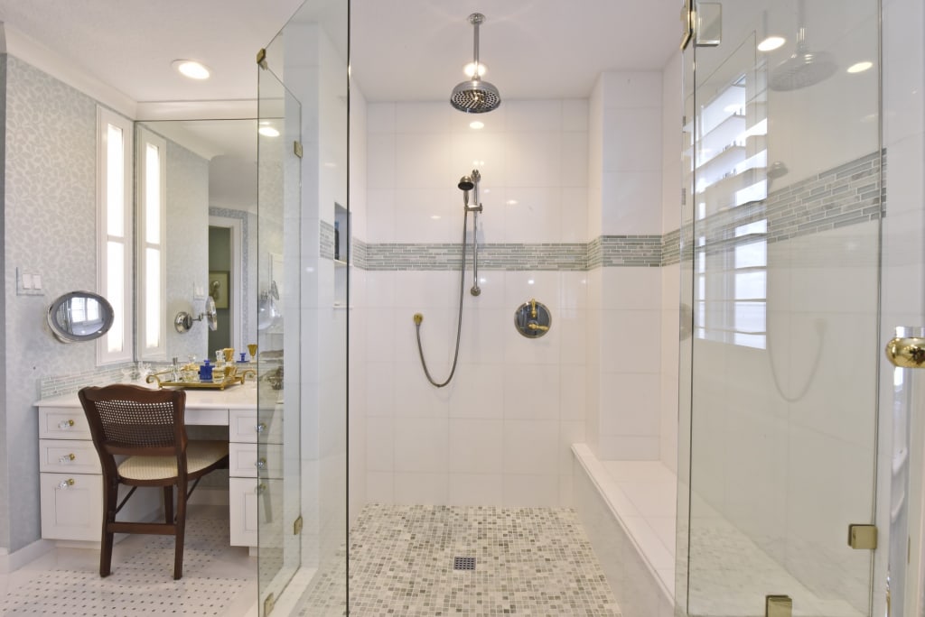 Modern bathroom with a large glass-enclosed shower, rain showerhead, hand-held shower, built-in bench, mosaic tile floor, and a vanity area with a chair, mirror, and various toiletries on the left.