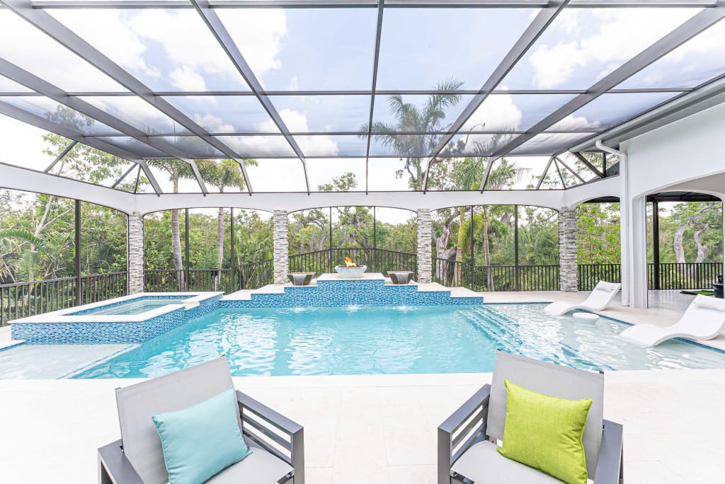 A luxurious screened-in outdoor pool area with a hot tub, lounge chairs in the water, palm trees, and two gray chairs with green and blue pillows on a white patio. The sky and trees are visible beyond the enclosure.