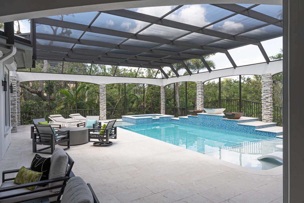 A screened-in outdoor pool area with lounge chairs, tables, and modern decor. The pool features a waterfall edge and two elevated planters, surrounded by tropical plants and trees visible through the screen enclosure.