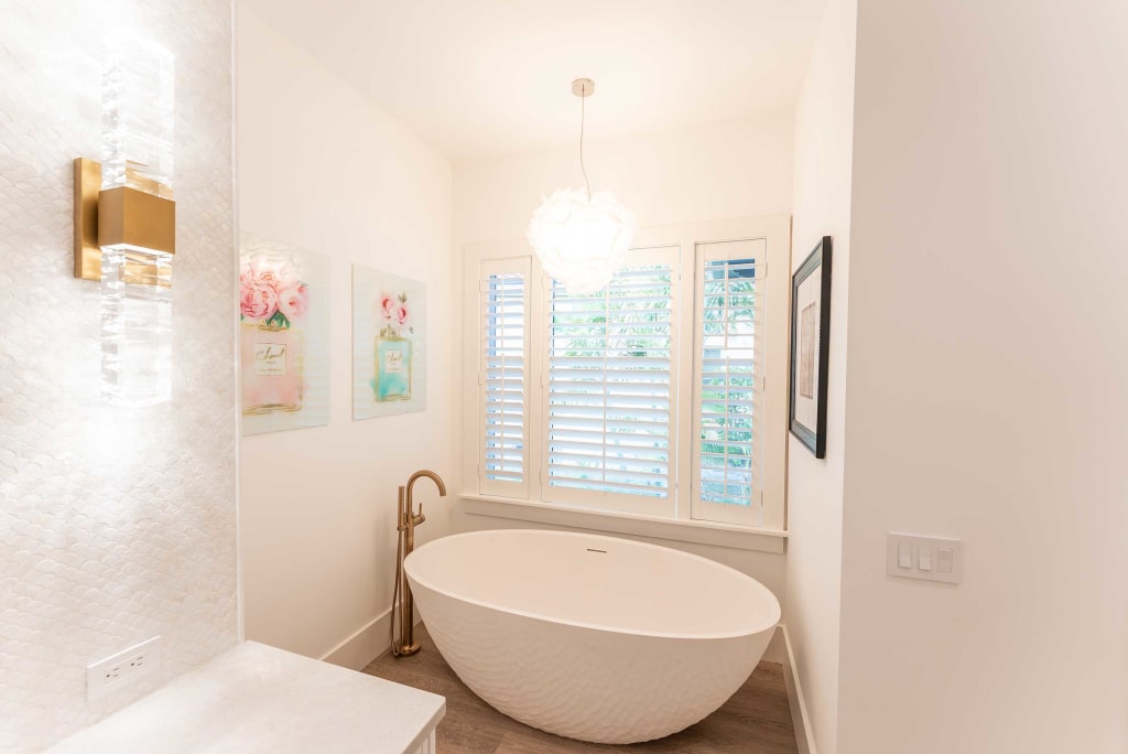 A modern bathroom with a white oval freestanding bathtub beneath a window with white shutters, a gold floor-mounted faucet, wall art with floral designs, and a pendant light hanging from the ceiling.