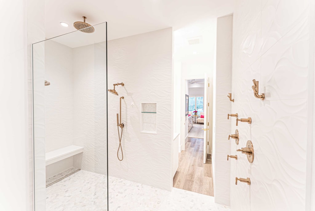 A modern, spacious shower with white textured walls, a built-in bench, multiple gold shower fixtures, and a glass partition. The shower opens to a hallway with wood flooring and a view into a living area.
