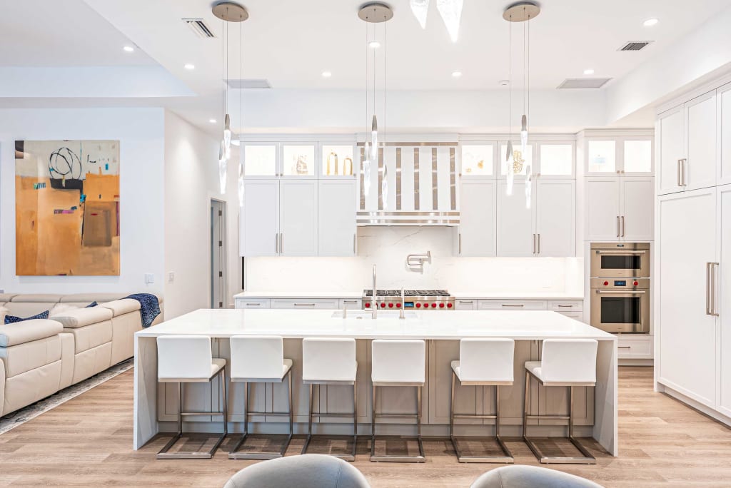 Modern white kitchen with a large island, five white bar stools, pendant lights, stainless steel appliances, and minimalist decor; a living area with abstract art is visible on the left.