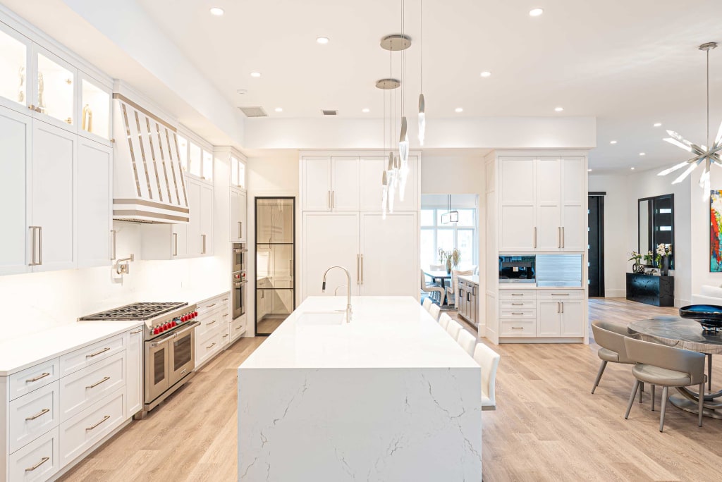 Modern kitchen with white cabinets, marble island with sink, stainless steel appliances, light wood flooring, pendant lights, and a dining area with round table and gray chairs. Bright, spacious, and open design.