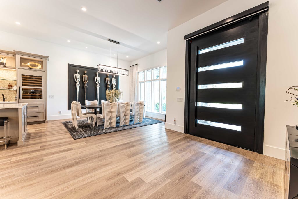 Modern dining room with a black front door, light wood flooring, a dining table with white chairs, contemporary chandelier, wall art of three metallic sculptures, and large windows letting in natural light.