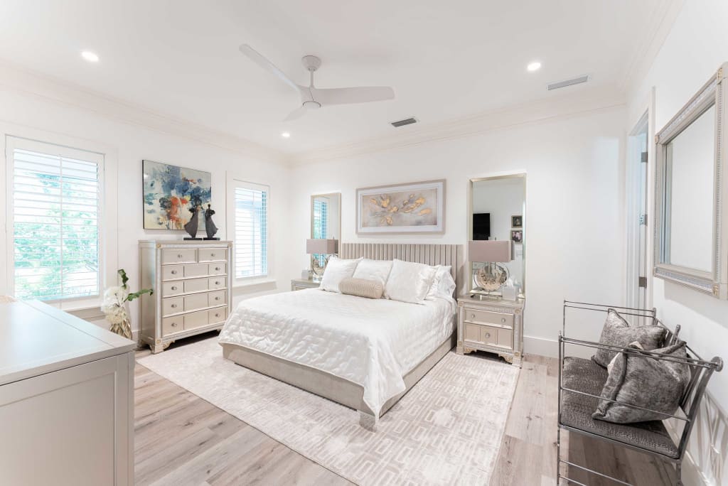 Bright, modern bedroom with a white bed, matching nightstands, dresser, and artwork on the walls. The room features light wood flooring, large windows with shutters, a ceiling fan, and neutral decor accents.