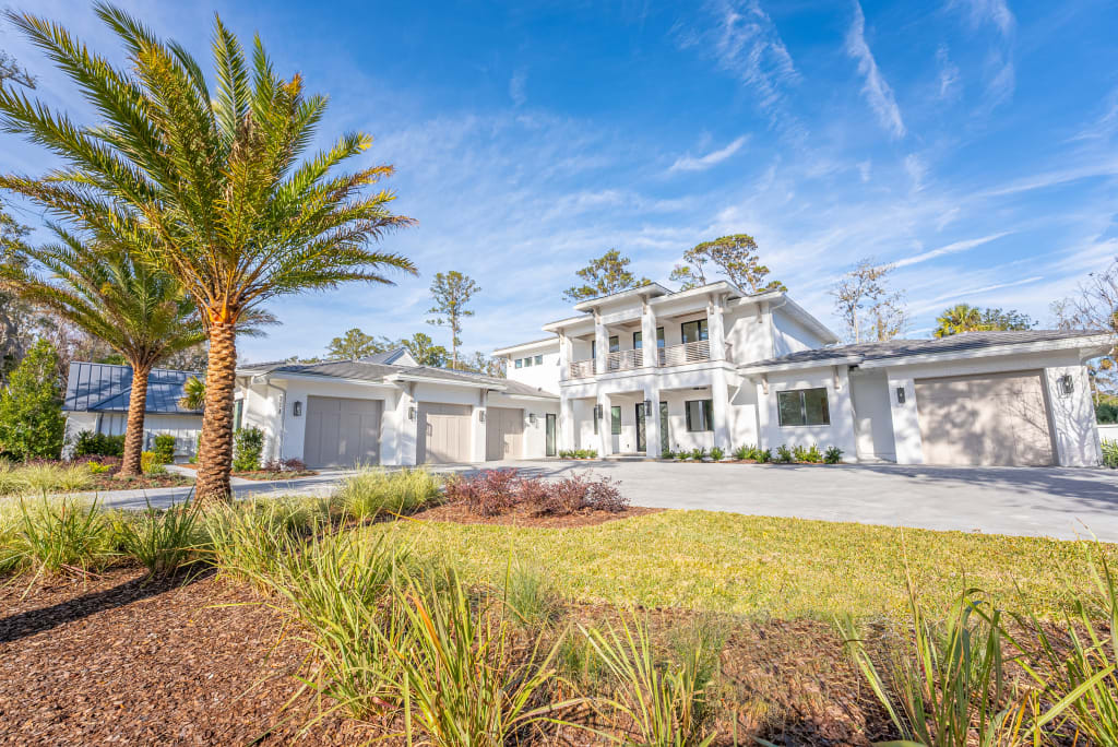 A large, modern white house with multiple garages, a manicured lawn, palm trees, and clear blue sky in the background—perfectly capturing the upscale lifestyle of homes in Nocatee FL.