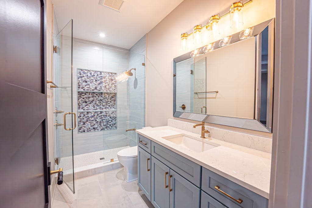 Modern bathroom in homes in Nocatee FL with a glass-enclosed shower, toilet, double-sink vanity with gold fixtures, large mirror, under-cabinet storage, and wall-mounted lights above the mirror.