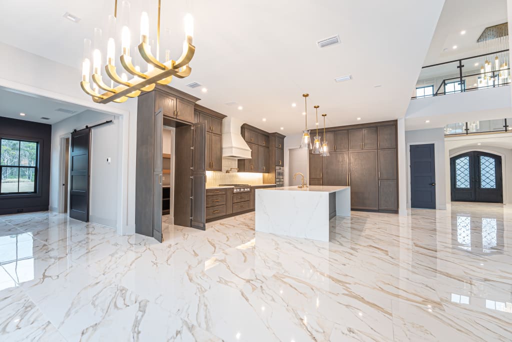Spacious modern kitchen with marble flooring, dark wood cabinets, a large island with a marble finish, gold chandeliers, and high ceilings—perfect for those seeking luxury in homes in Nocatee FL. Multiple doorways and large windows let in natural light.