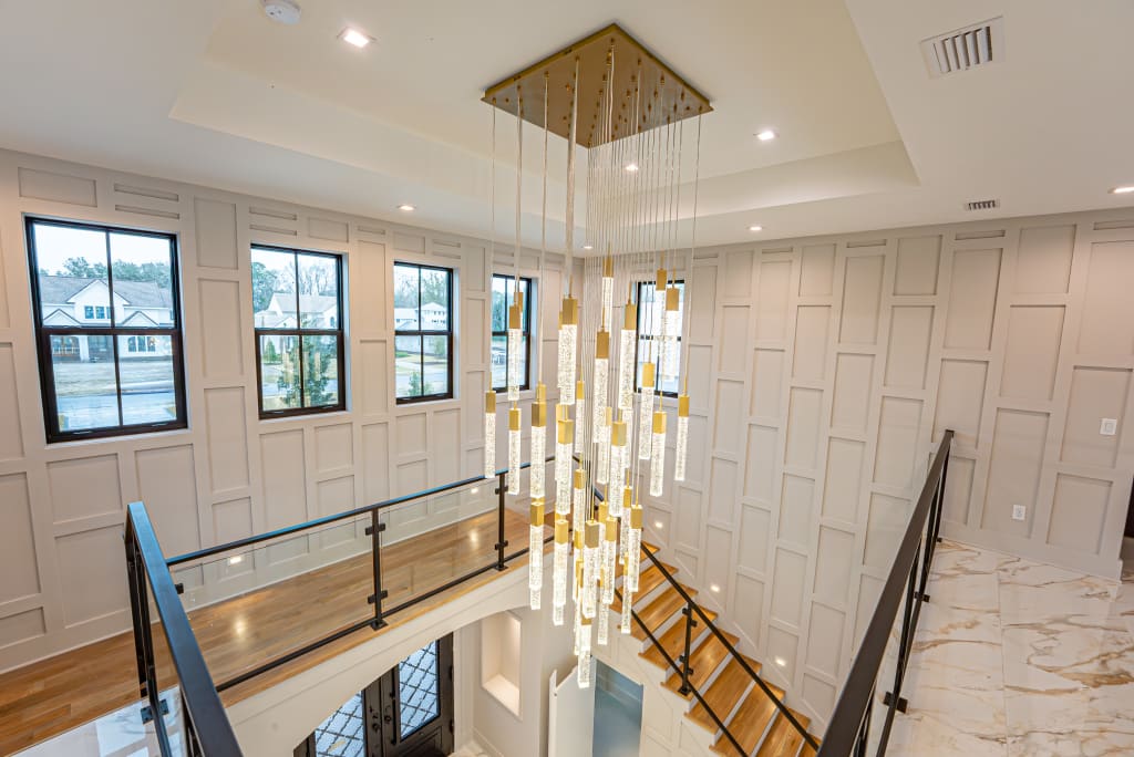 A modern chandelier with hanging glass rods is suspended in a bright, double-height foyer found in many homes in Nocatee FL, featuring geometric wall paneling, large windows, a glass railing, and a staircase with wooden steps.