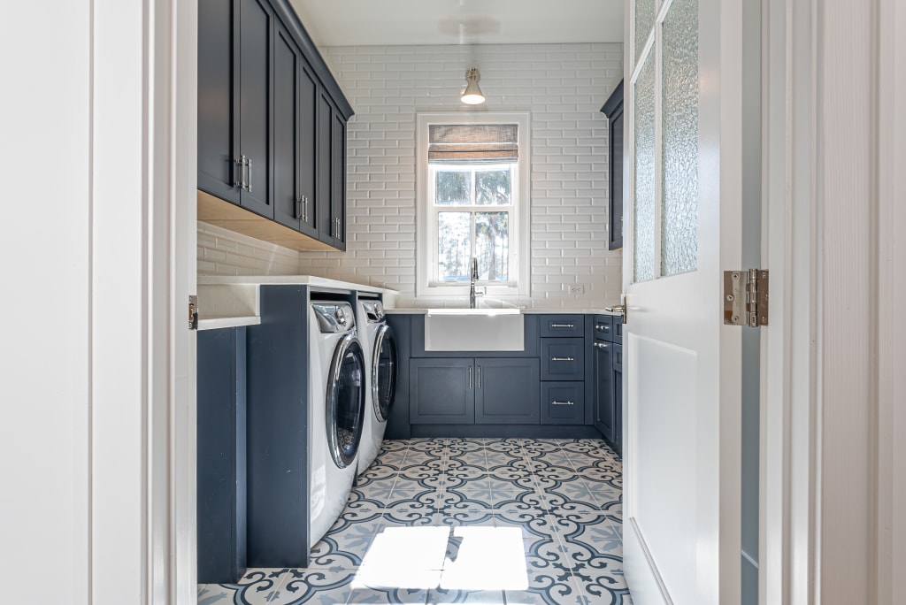 A modern laundry room with dark blue cabinets, a farmhouse sink under a window, white subway tile walls, and patterned blue and white floor tiles offers stylish utility in many homes in Nocatee FL.