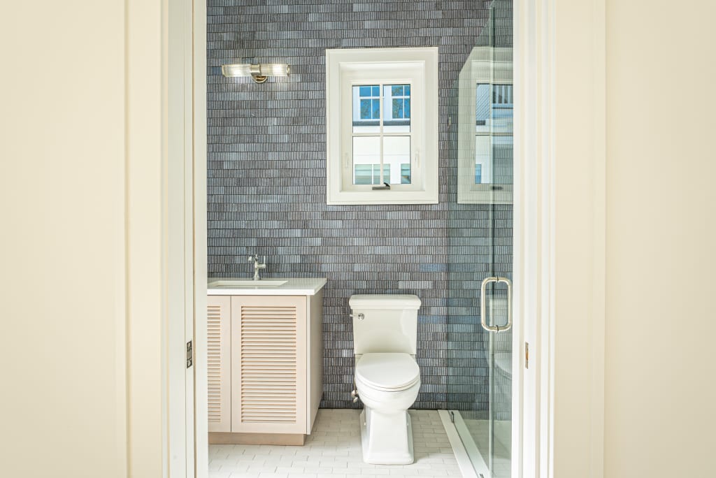 A bright, modern bathroom in homes in Nocatee FL features a white toilet, a glass shower, a single-sink vanity with wooden cabinet doors, and a window above—all set against a wall of gray rectangular tiles.