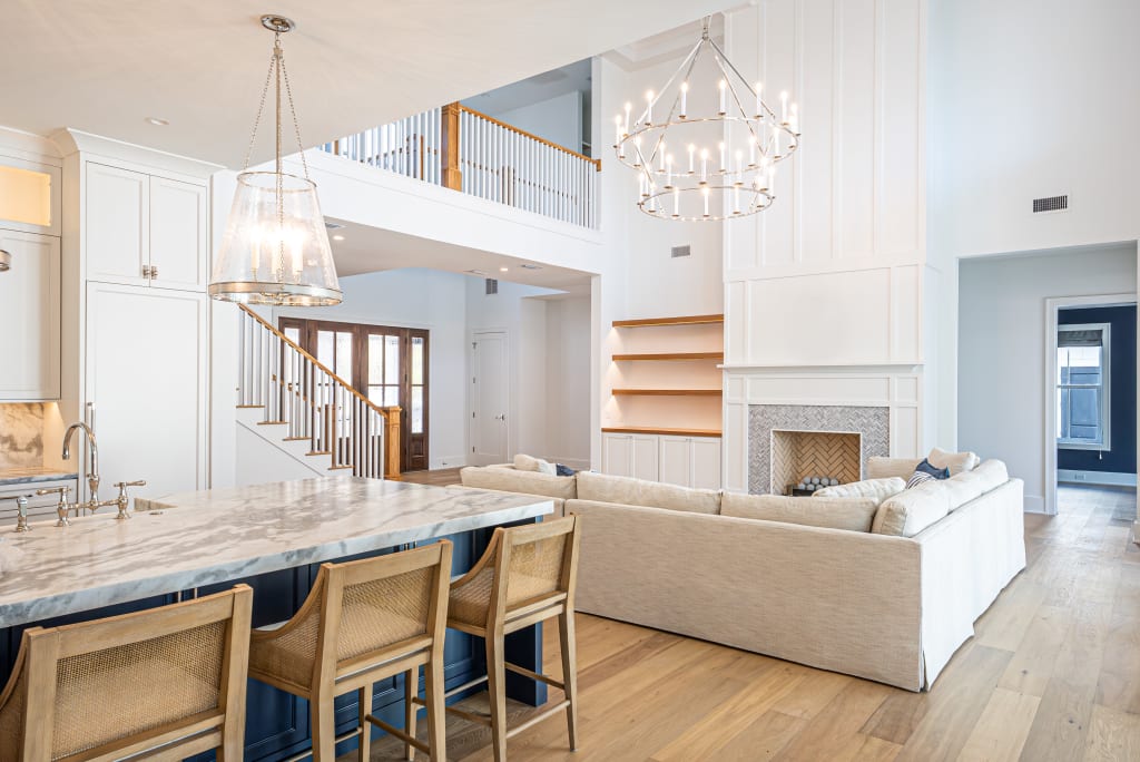 Spacious, modern living room and kitchen in one of the stunning homes in Nocatee FL, featuring a large sectional sofa, marble island with wooden barstools, chandeliers, fireplace, built-in shelves, hardwood floors, and an open staircase with balcony railing.