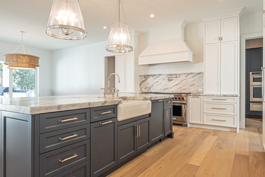 Modern kitchen in Homes in Nocatee FL featuring a large dark blue island, marble countertops, farmhouse sink, white cabinets, stainless steel appliances, and wooden floors. Two glass pendant lights hang above the island.