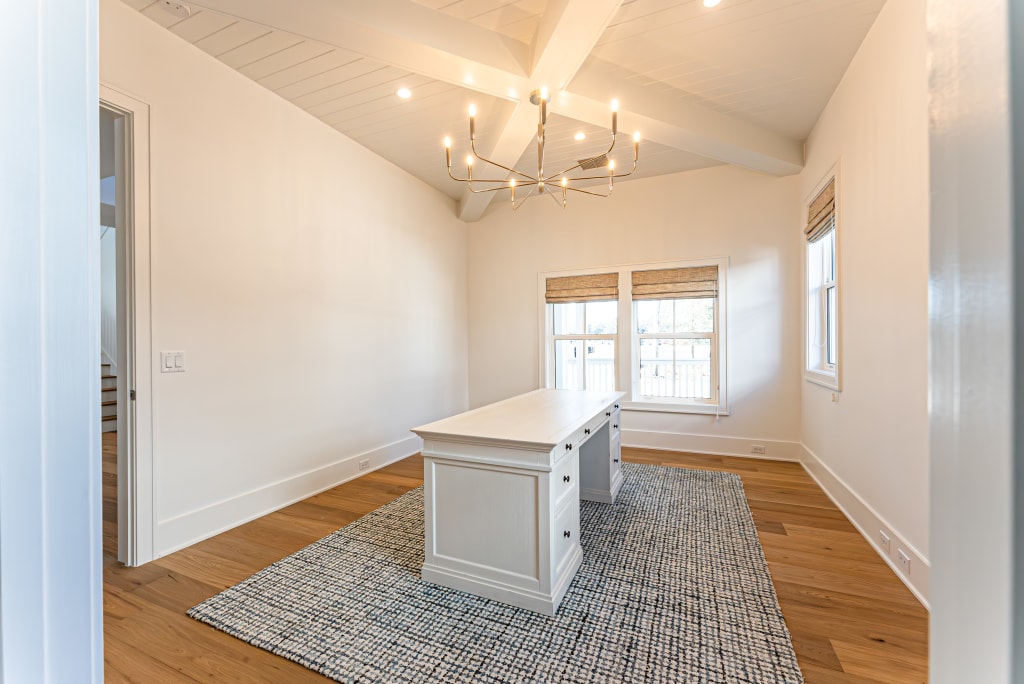 A bright home office with a white desk, a textured rug, wooden floors, large windows with shades, and a modern chandelier on a high beamed ceiling—reflecting the spacious, minimal style found in many homes in Nocatee FL.