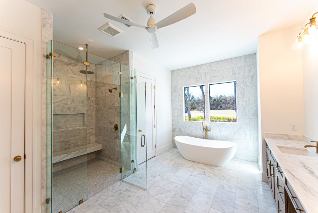 Bright, modern bathroom with marble floors and walls, glass shower with a bench, freestanding bathtub by two windows, double vanity with sinks, and a ceiling fan—typical of luxury homes in Nocatee FL.