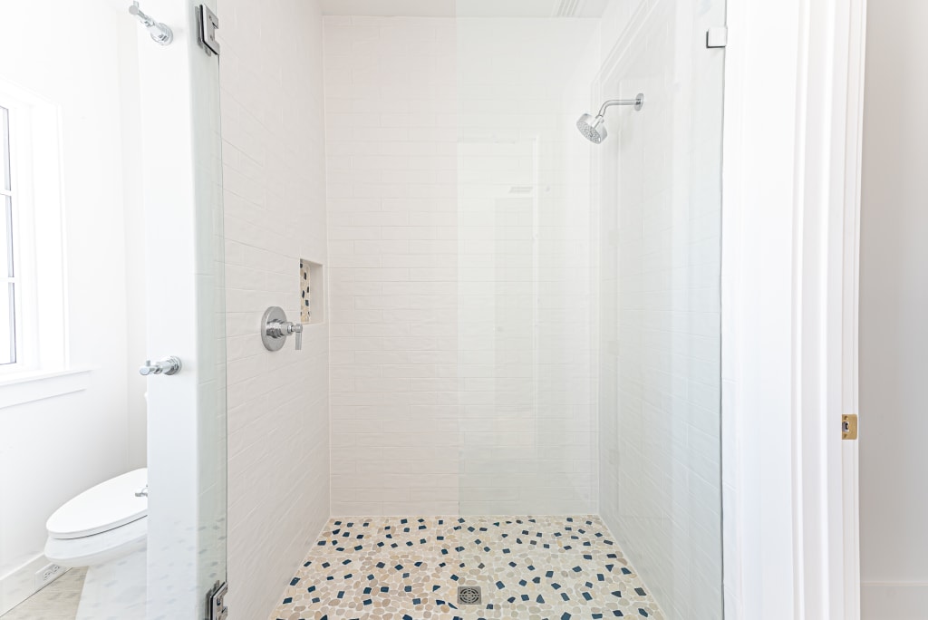 A bright, modern walk-in shower with a glass door, white tiled walls, built-in shelf, silver fixtures, and a pebble-patterned floor—designed to reflect the elegant style found in many homes in Nocatee FL. A toilet is visible to the left outside the shower.