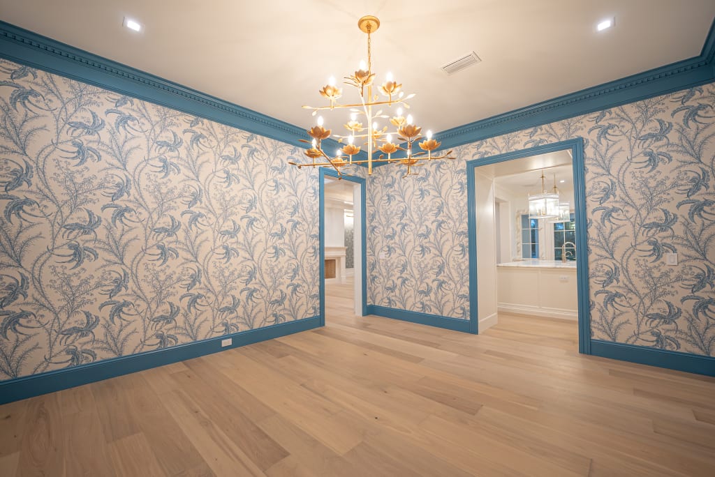 A bright, empty room with light wood floors, blue floral wallpaper, and blue trim. A modern gold chandelier hangs from the ceiling. Perfect for showcasing elegant Homes in Nocatee FL, with two doorways leading to adjoining rooms.