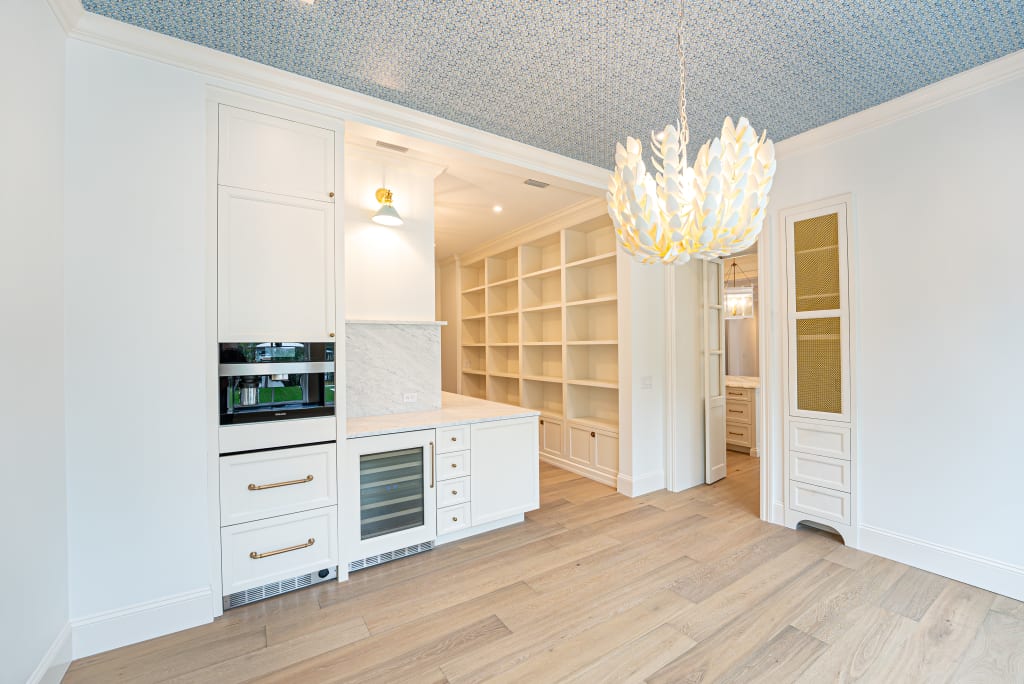Modern pantry with white cabinetry, built-in oven, wine fridge, open shelving, light wood floors, and a patterned blue ceiling. Perfect for homes in Nocatee FL, it features a decorative white pendant lamp and visible hallway with extra shelving.
