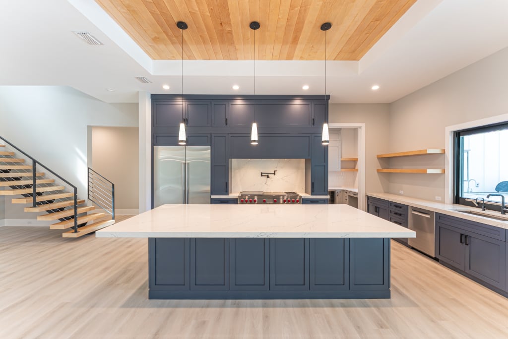 Modern kitchen with a large marble island, navy cabinets, pendant lights, light wood floors, open shelves, stainless steel appliances, and a wooden ceiling accent above the island—perfect for stylish homes in Nocatee FL. A staircase is visible on the left.